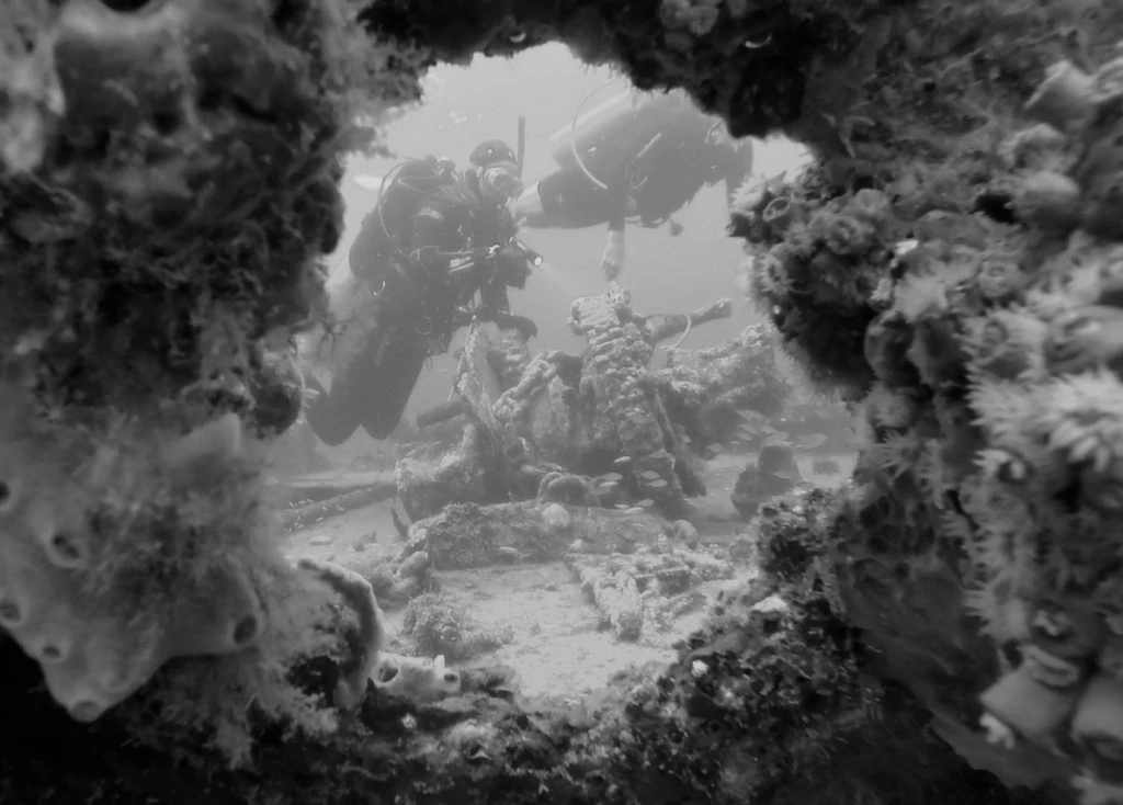 black-and-white 'through the porthole' image of divers on a wreck.